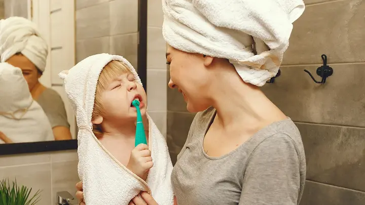 A woman in a hair towel holding a toddler who is brushing his teeth with issa 4 baby toothbrush